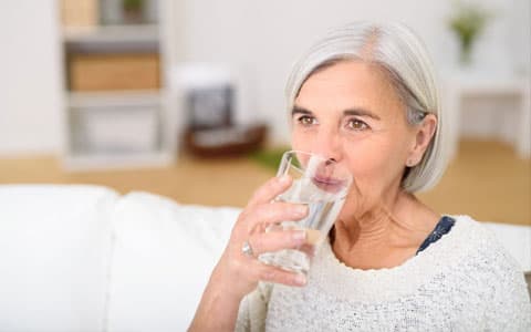 Elderly Woman Sat Down On Sofa Drinking Glass Of Water