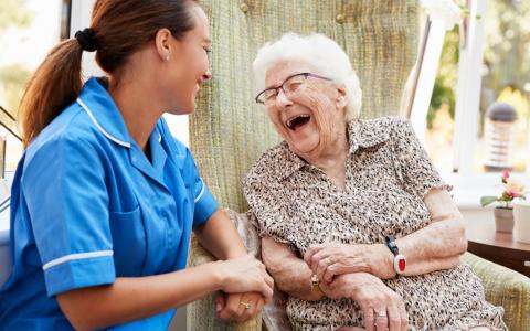 Young Carer Laughing With Elderly Woman Outside On Bench